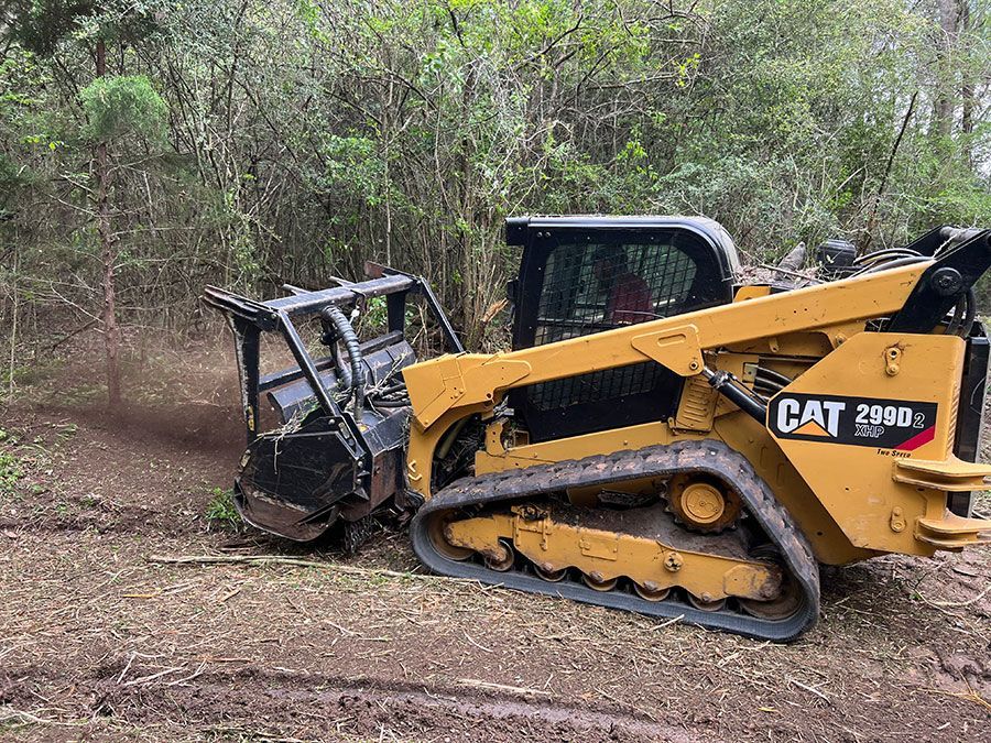 Yellow CAT skid steer pushing a black metal attachment through muddy woodland terrain