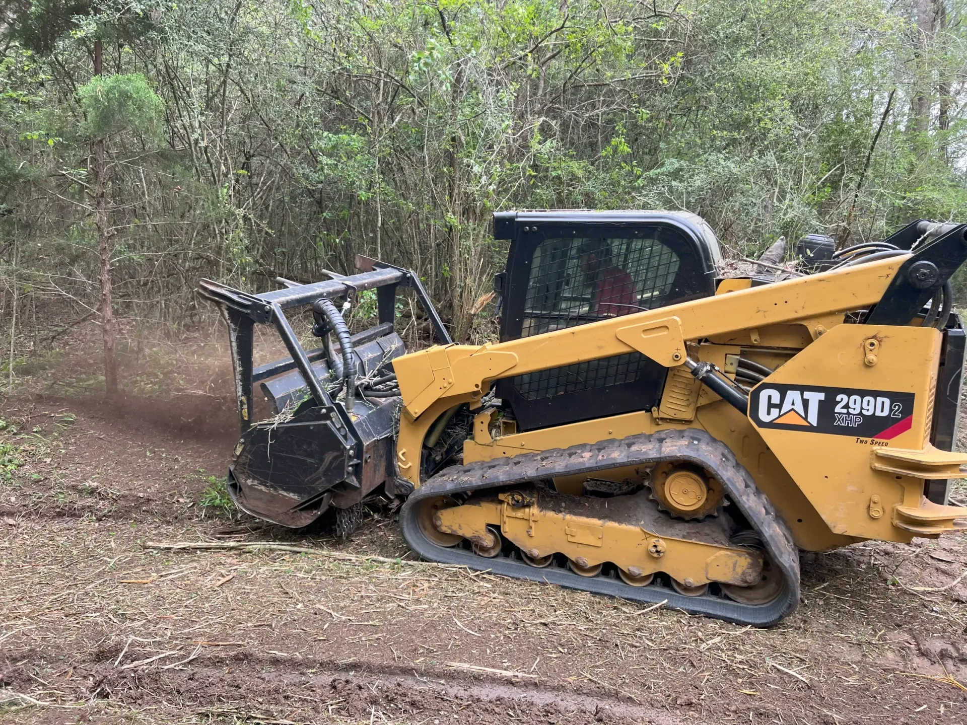 Yellow CAT skid steer clearing brush in a wooded area, kicking up dust.