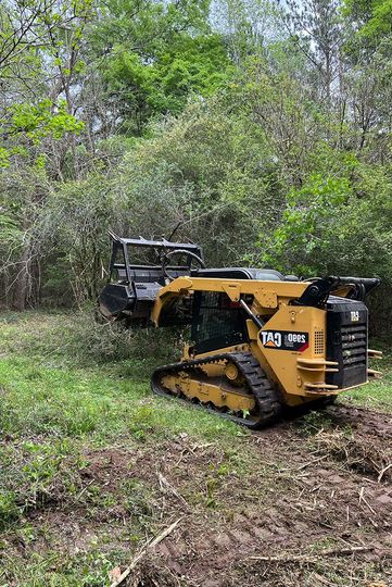 Yellow tracked skid steer working on a wooded path with a black trailer behind it