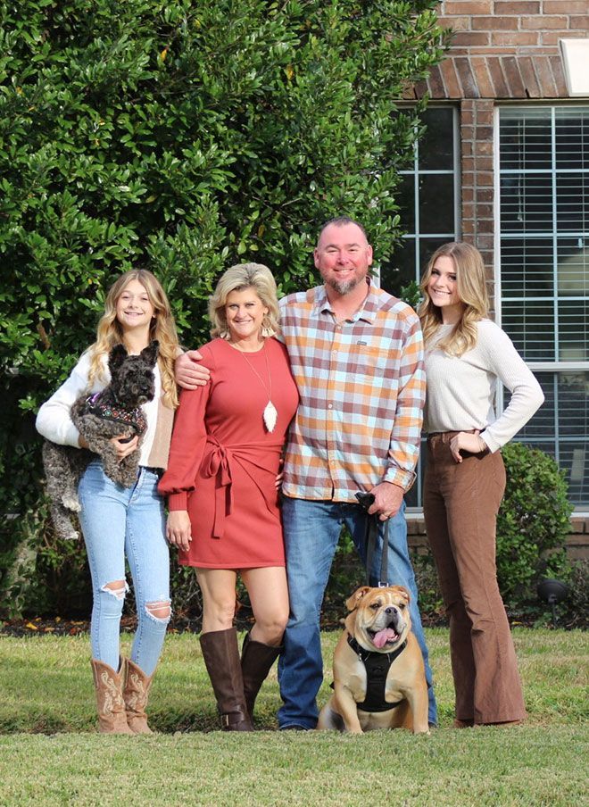 Four people posing outdoors in front of a house, with a cat and a dog in front.