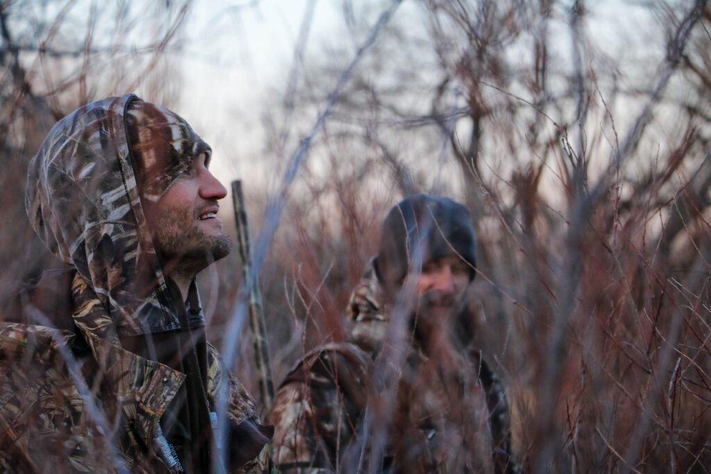 Two people in winter coats stand among tall dry grass in a foggy field