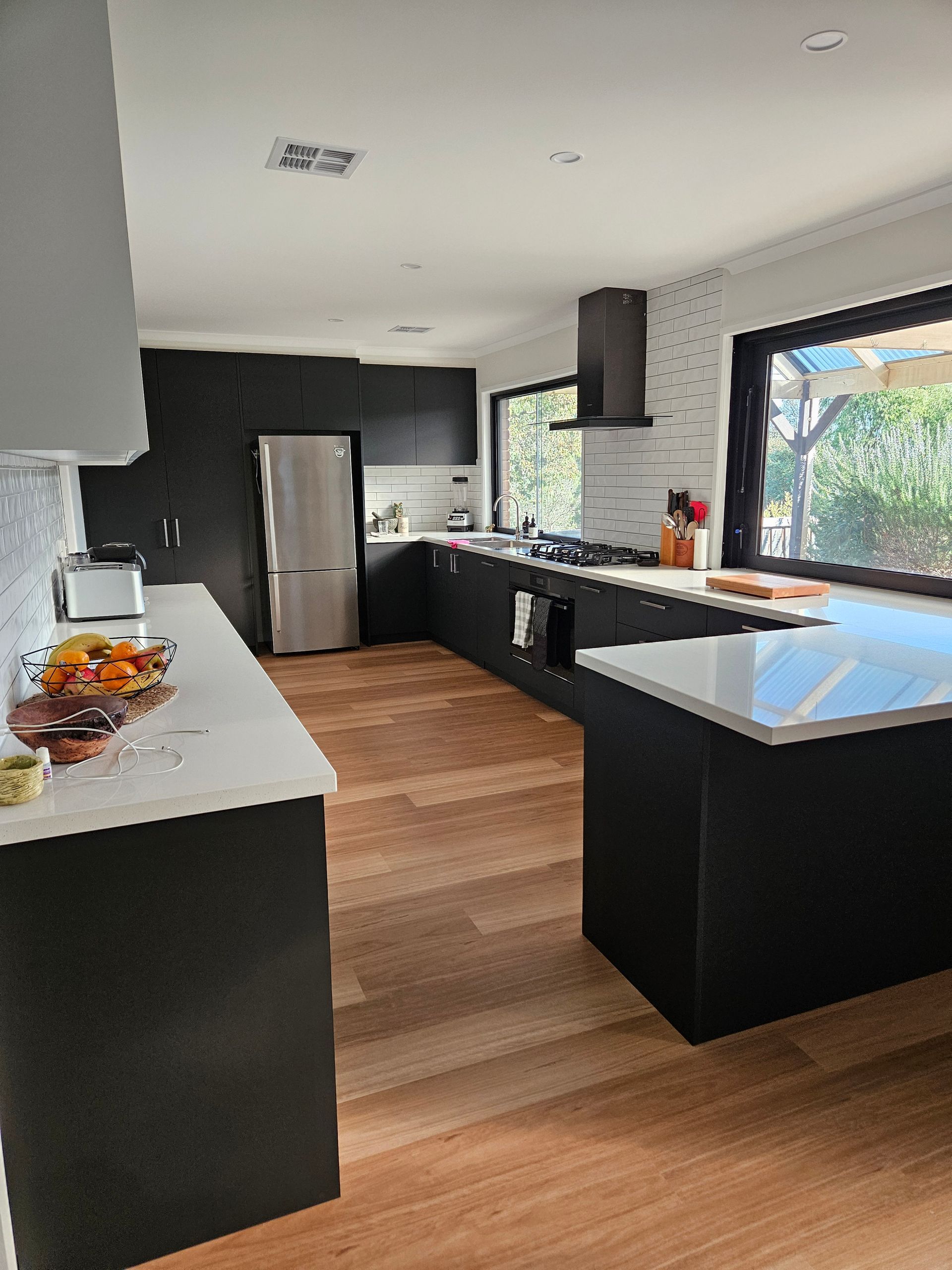 A kitchen with black cabinets and wooden floors and a stainless steel refrigerator.