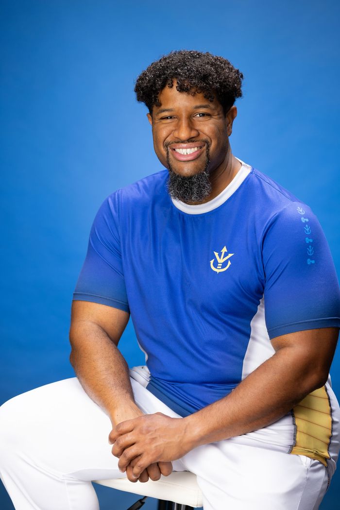 A man with a beard is sitting on a chair wearing a blue shirt and white pants.