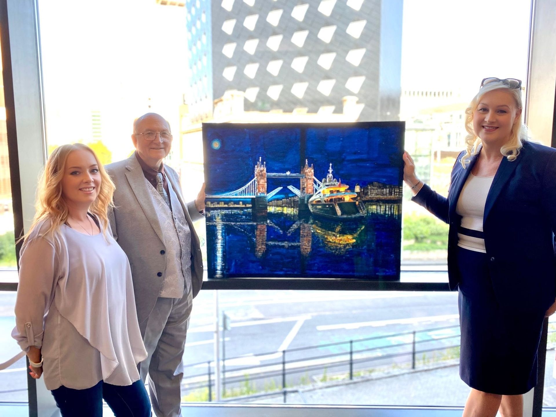Renowned Welsh Artist Jeffrey Phillips and International Agent Donna Phillips stand with a painting of a Superyacht at Tower Bridge, London. To the left is Kerry Ballard, CEO at Superyacht Sales UK