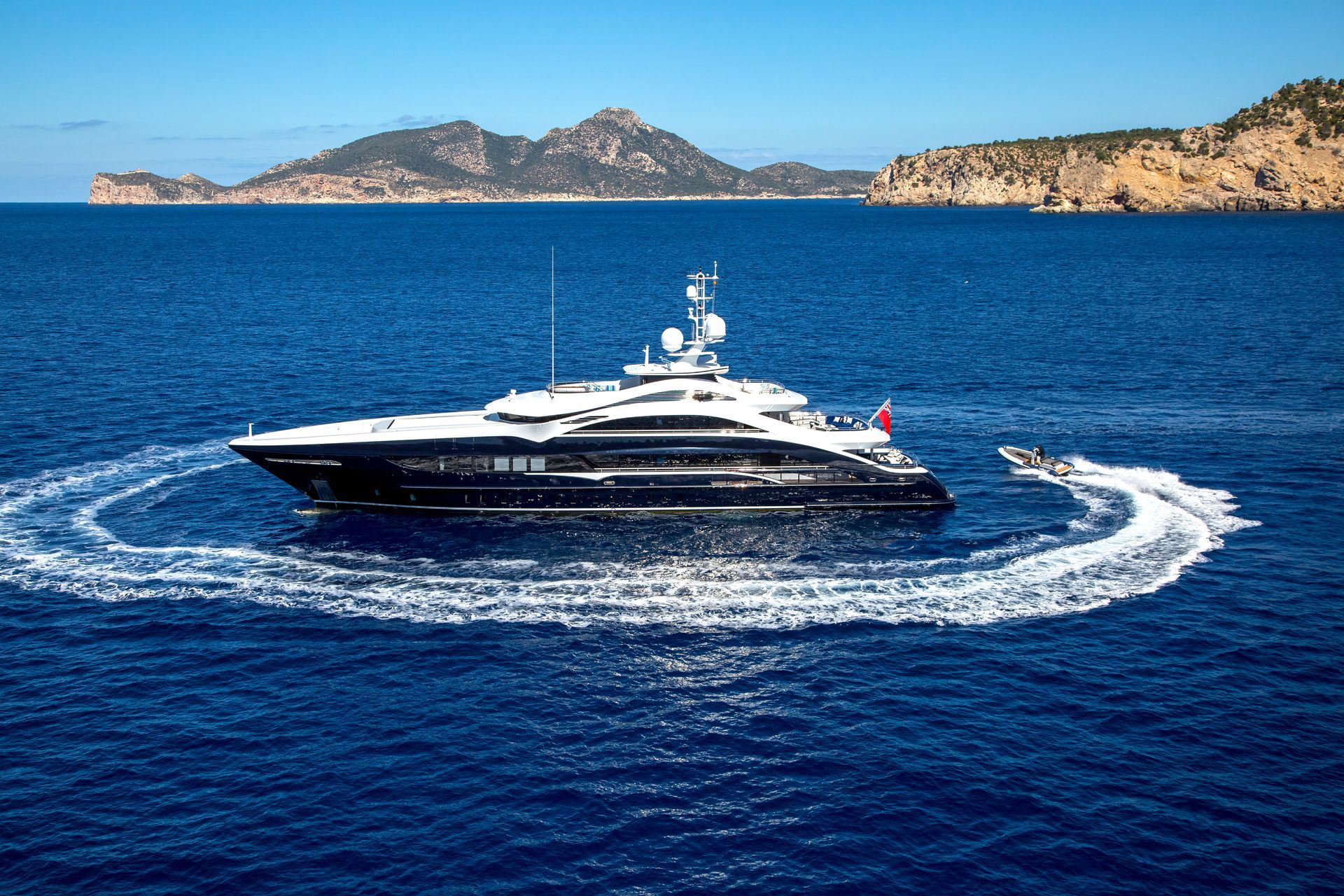 Superyacht with blue hull being circled by jetski off a mediterranean coast