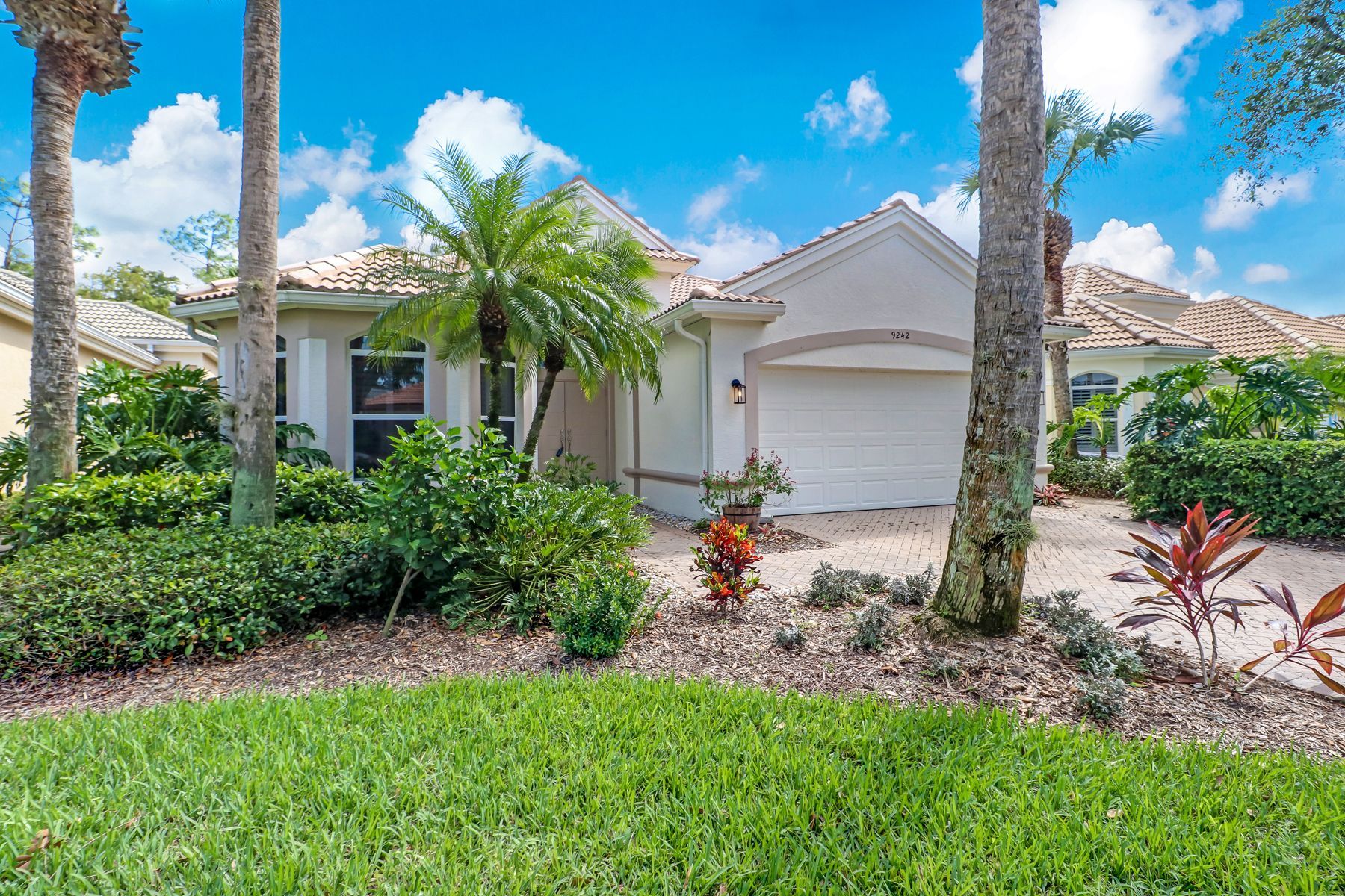 Single-story white house with garage, palm trees, and lush green lawn under a bright blue sky