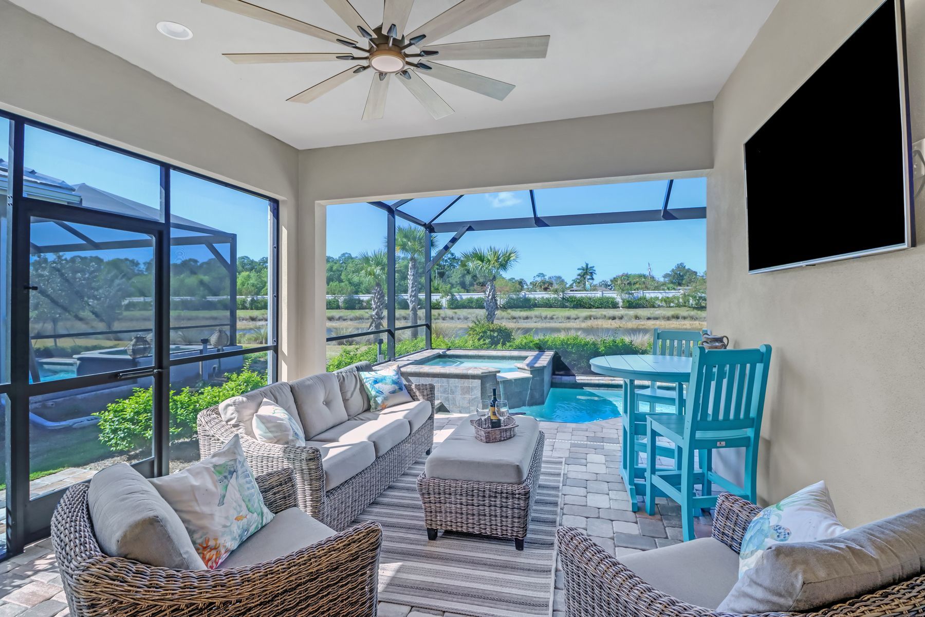 Covered patio lounge with white wicker seating, turquoise chairs, and a pool view through large windows