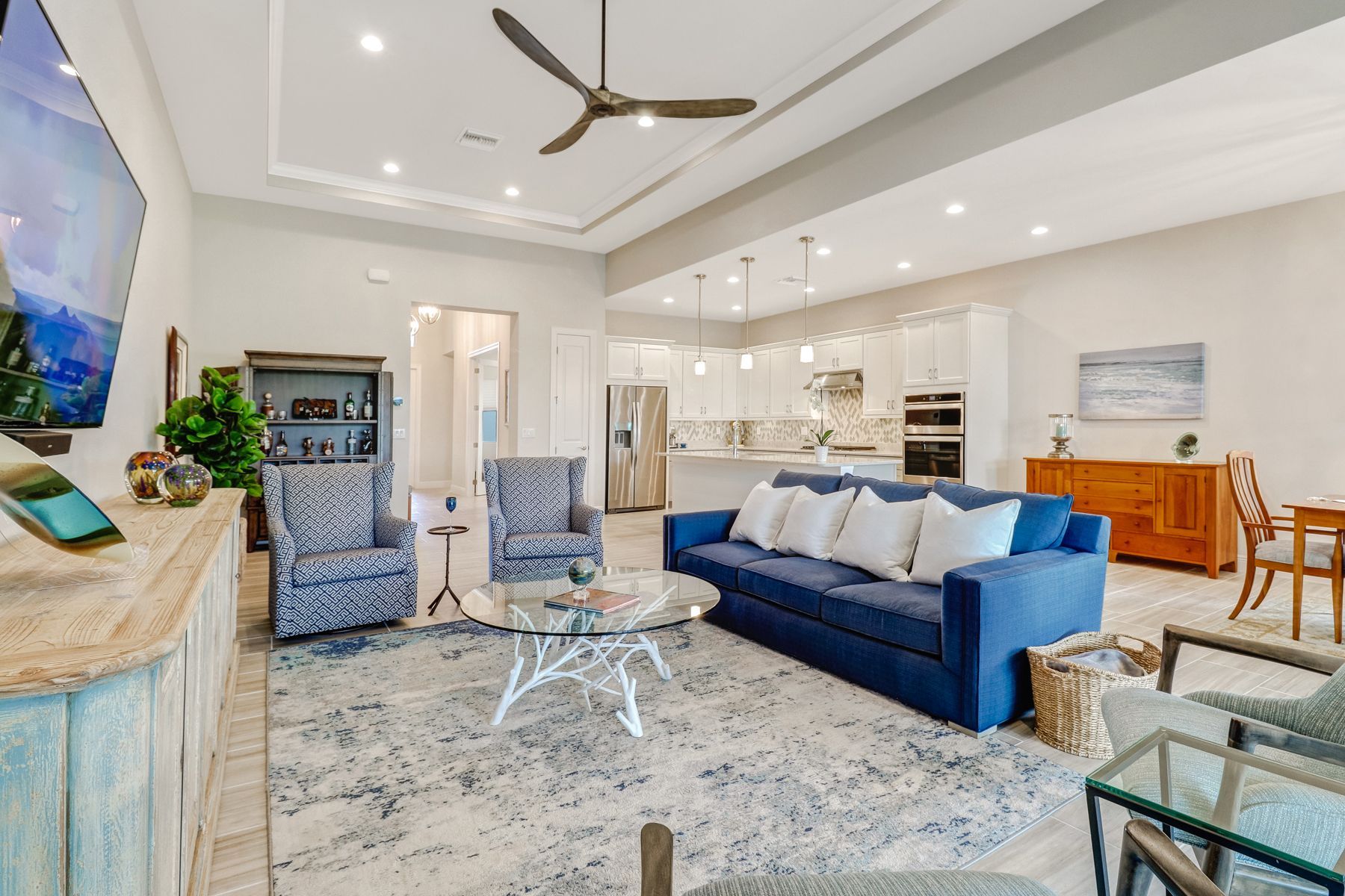 Bright modern living room with blue sofa, patterned chairs, glass tables, and open kitchen