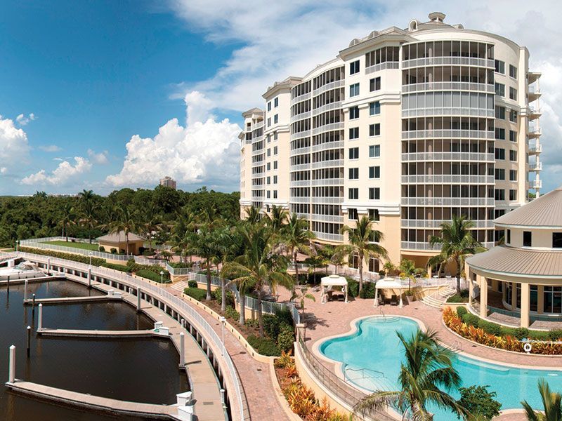 Waterfront resort with white high-rise, palm trees, pools, and marina under a blue sky
