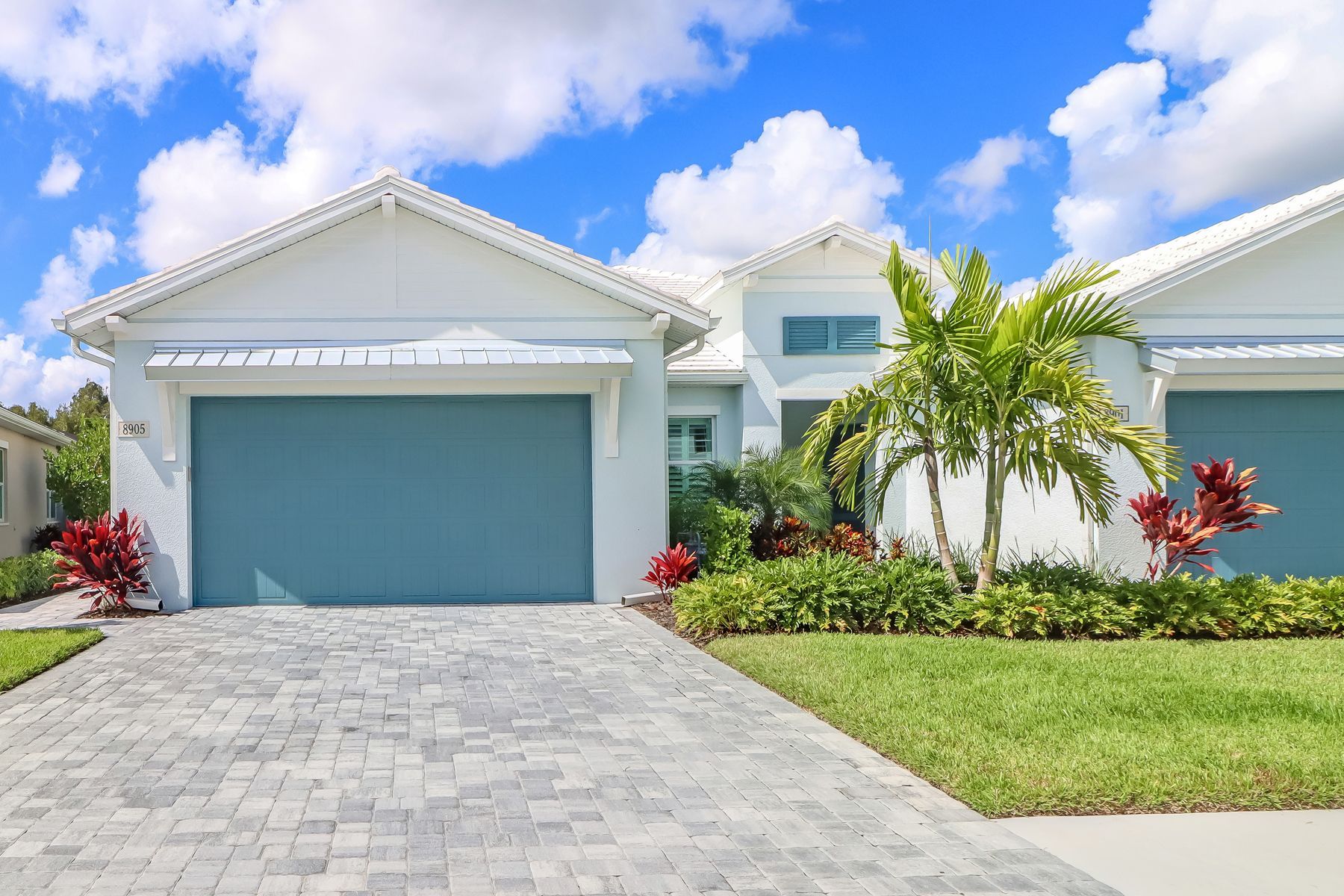 Single-story white house with blue garage door, palm tree, and stone driveway under a sunny sky