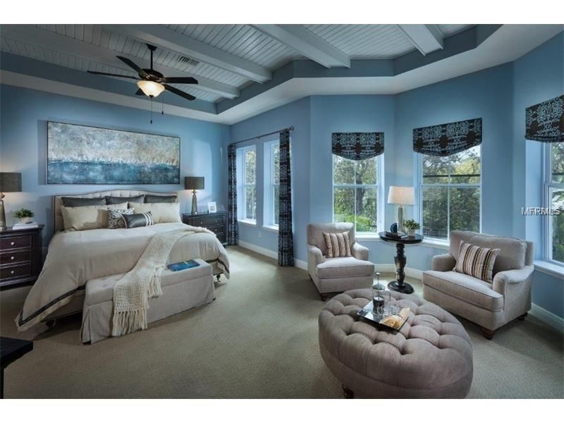 Blue bedroom with a bed, two armchairs, a tufted ottoman, and large windows overlooking water.