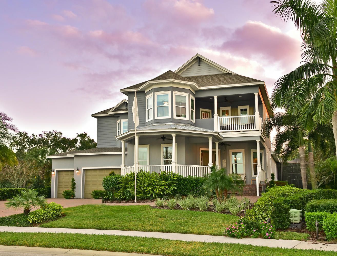 Two-story suburban house with a garage, wraparound porch, and lush landscaping at sunset