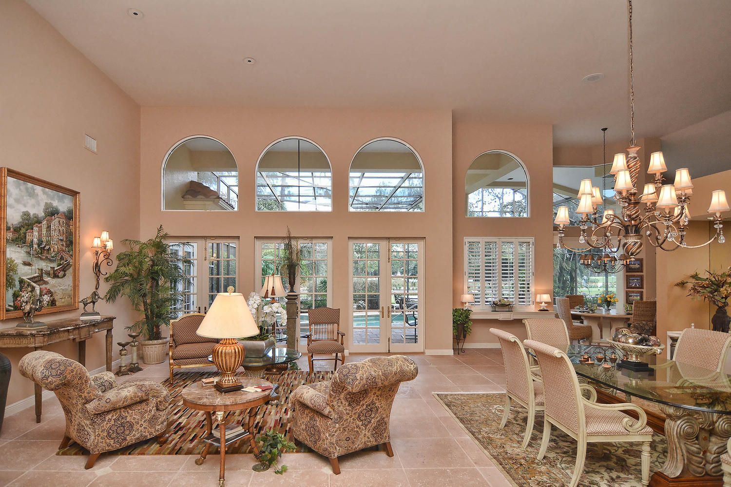 Elegant living room with arched windows, floral chairs, chandelier, and warm peach walls.