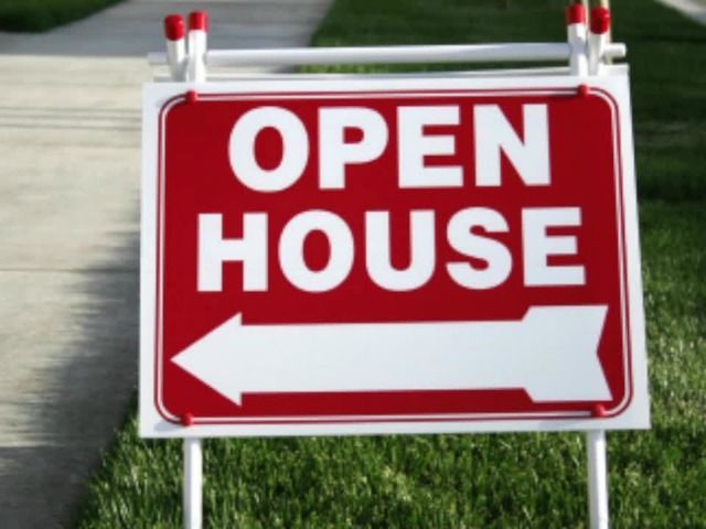 Red open house yard sign with white text and a left-pointing arrow on grass by a curb