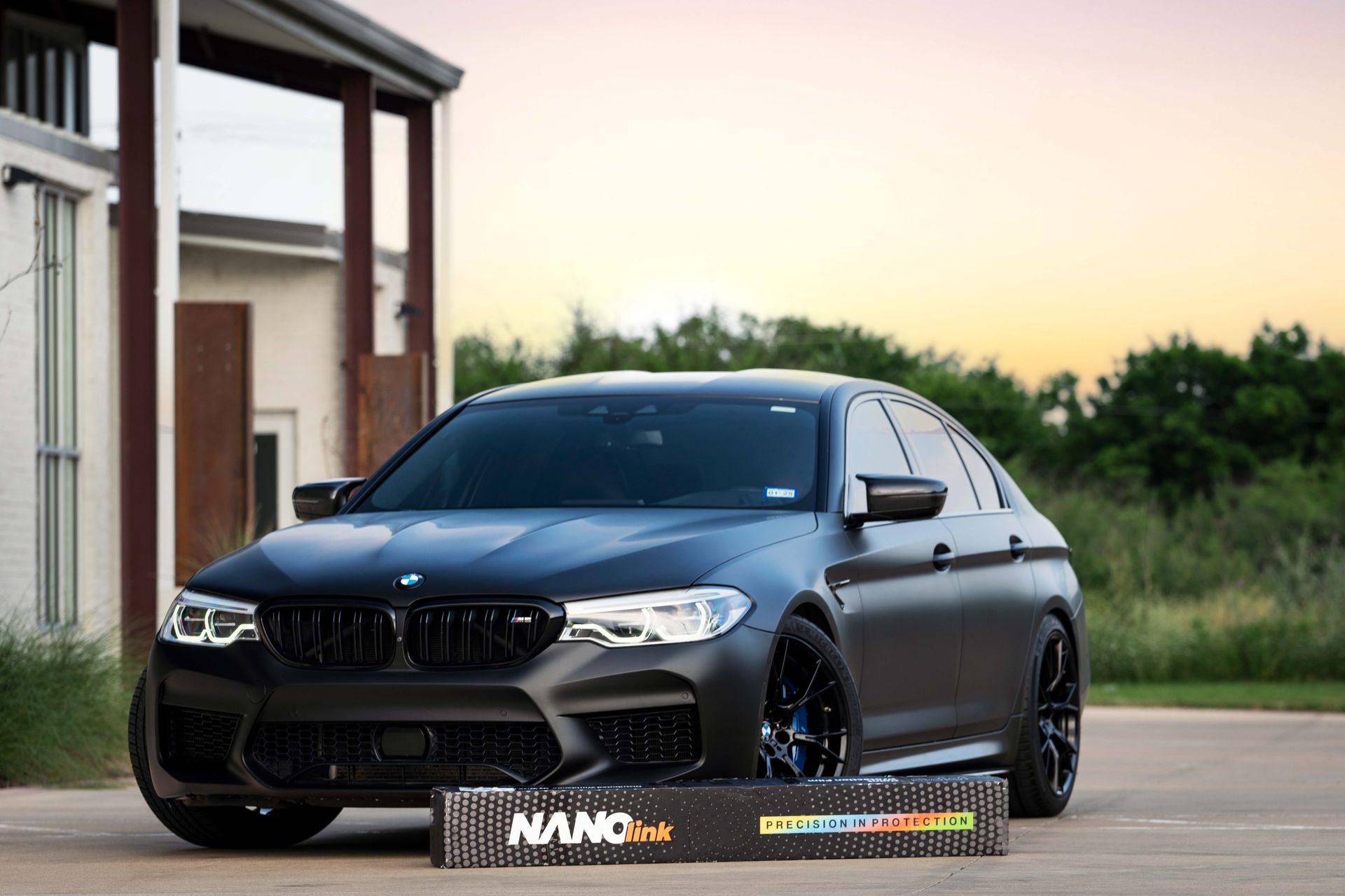 Satin black BMW M5 sedan parked outdoors with a roll of NanoPro film in front.