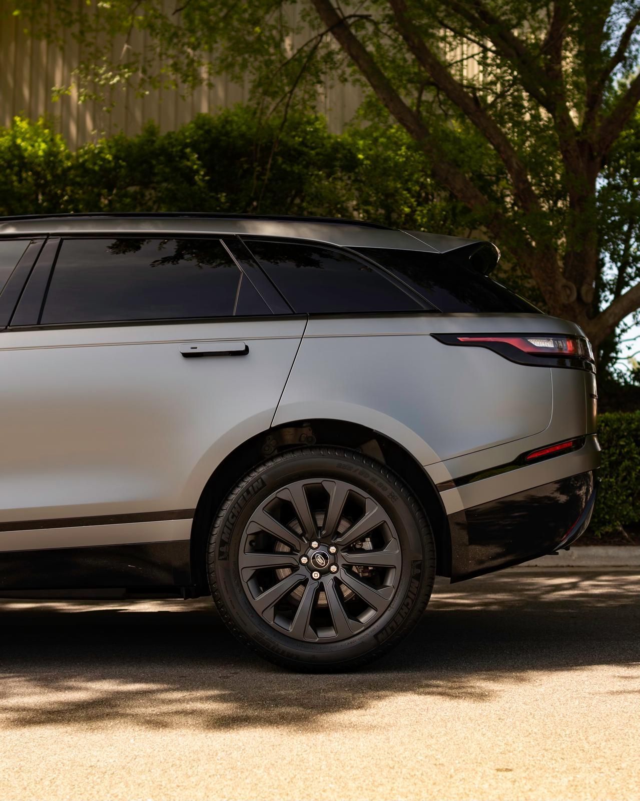 Silver Range Rover Velar SUV parked on pavement, dark wheels, black trim, trees and greenery in the background.