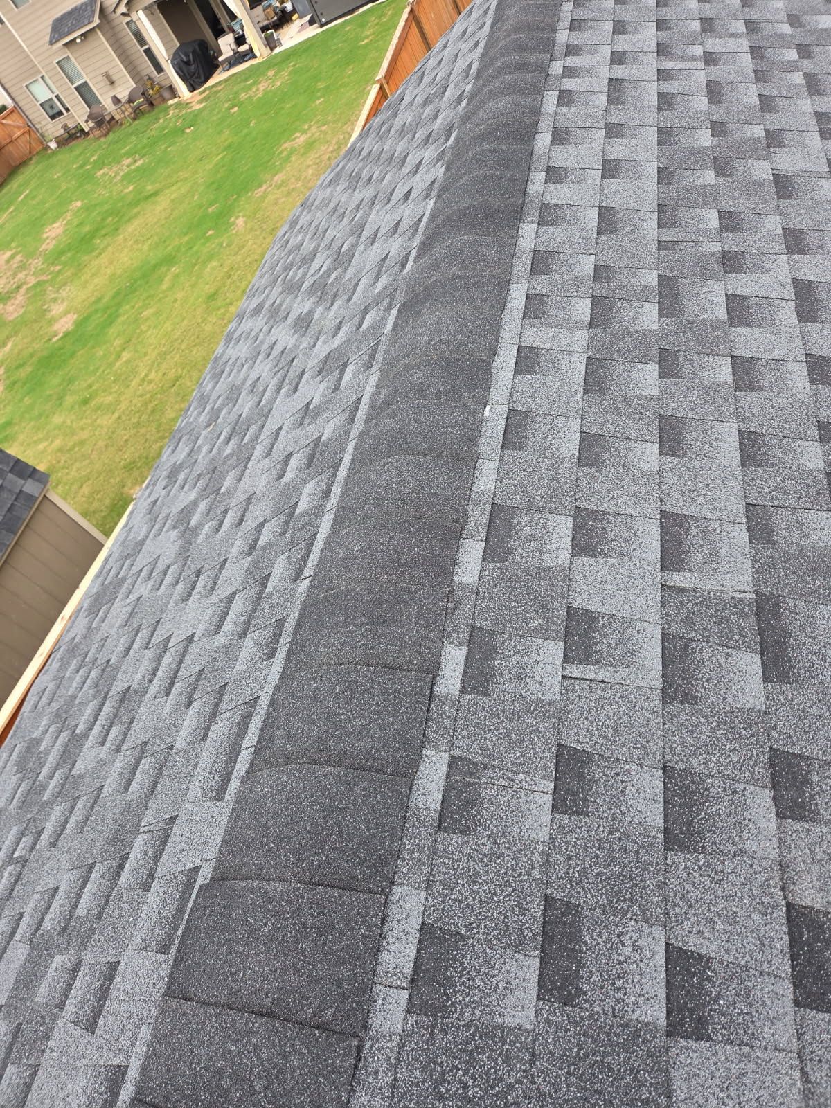 Gray asphalt shingle roof with a dark gray ridge cap, viewed from above, with green grass in the background.
