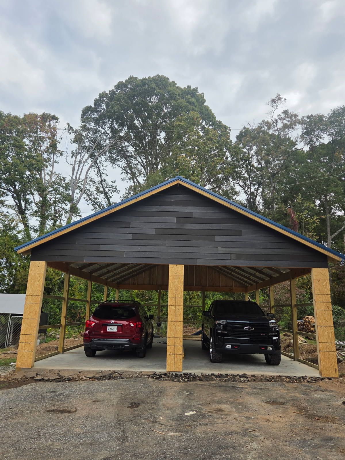 Two cars parked under a wooden carport with a dark gray facade, surrounded by trees.