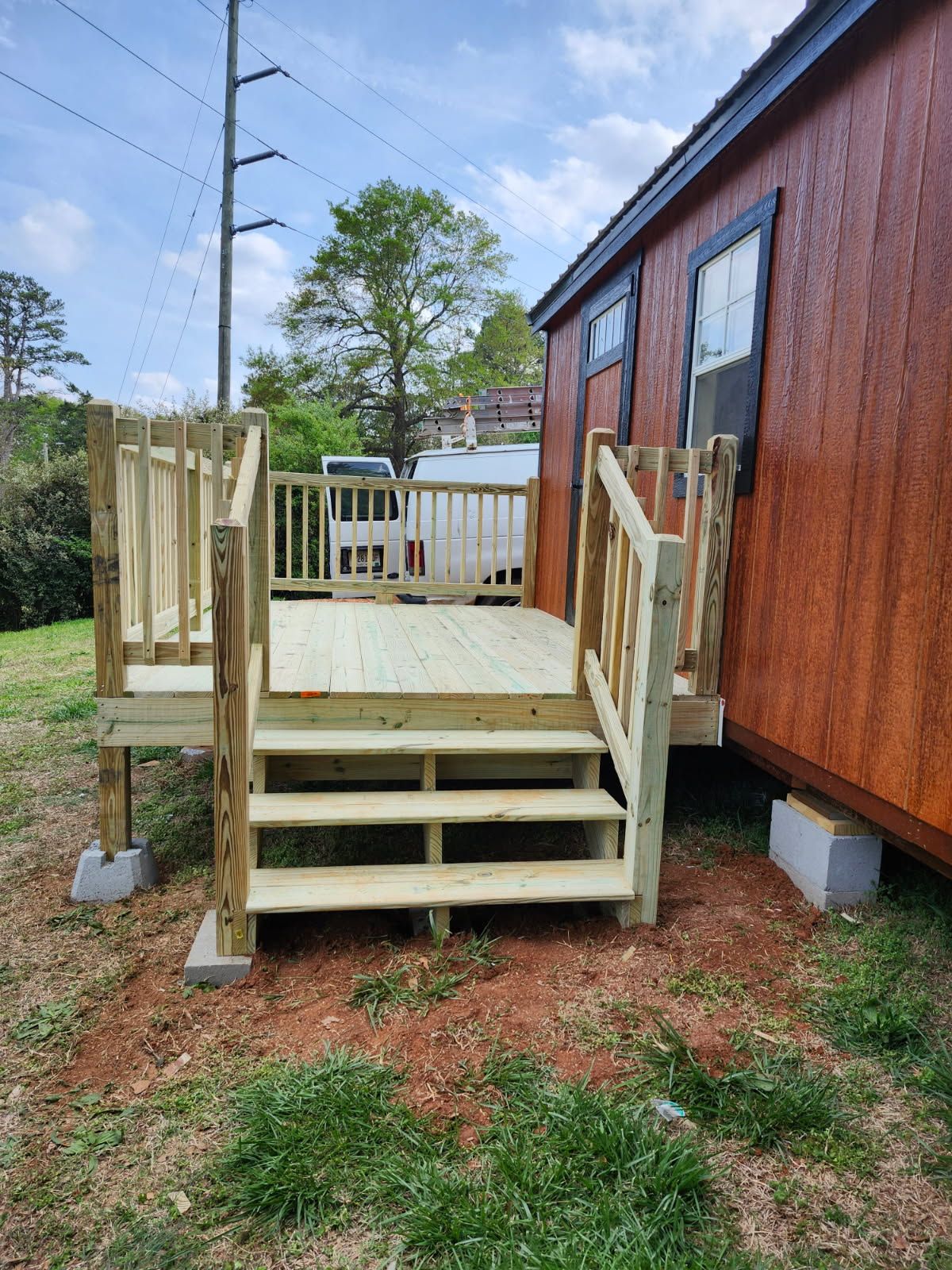 Wooden deck and stairs next to a reddish-brown structure, on concrete blocks, outdoors with cloudy sky.