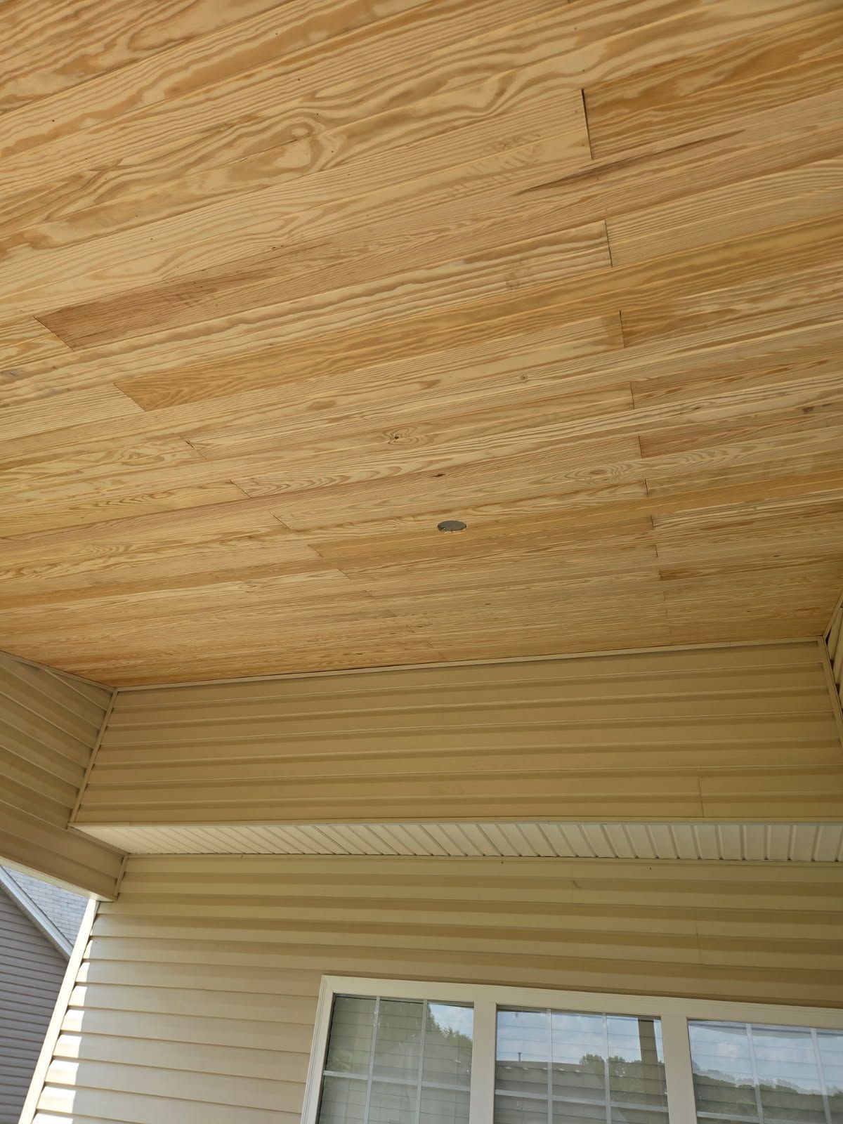Light wood plank ceiling above tan vinyl siding and windows.