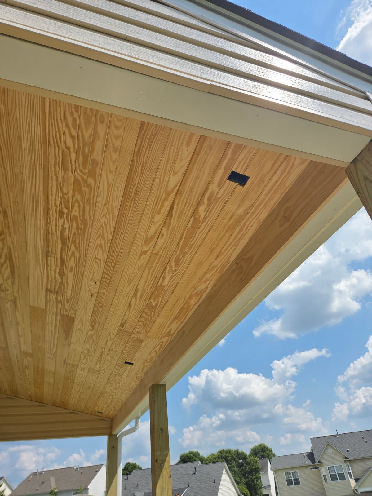 Wooden porch ceiling with light-colored planks, bordered by white trim. Blue sky and clouds visible.
