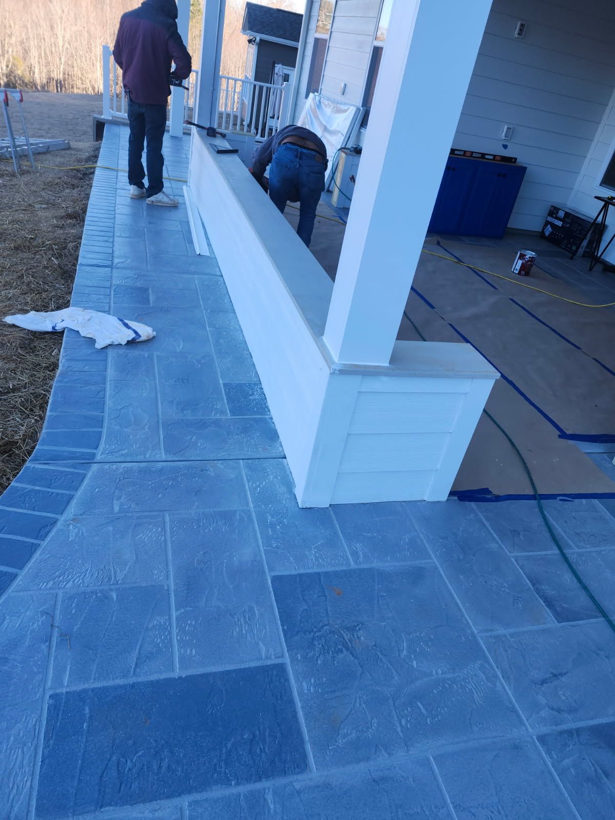 Workers installing white trim around a porch column, on a blue stone patio.