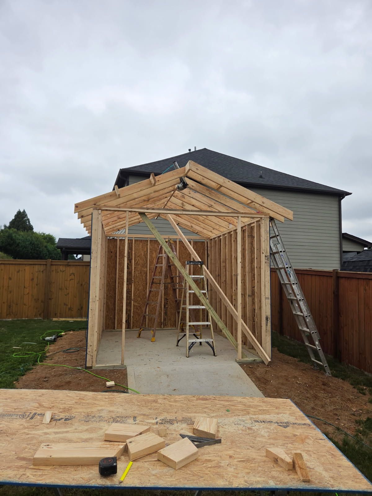 Wooden shed under construction in a backyard, ladders inside, cloudy sky.