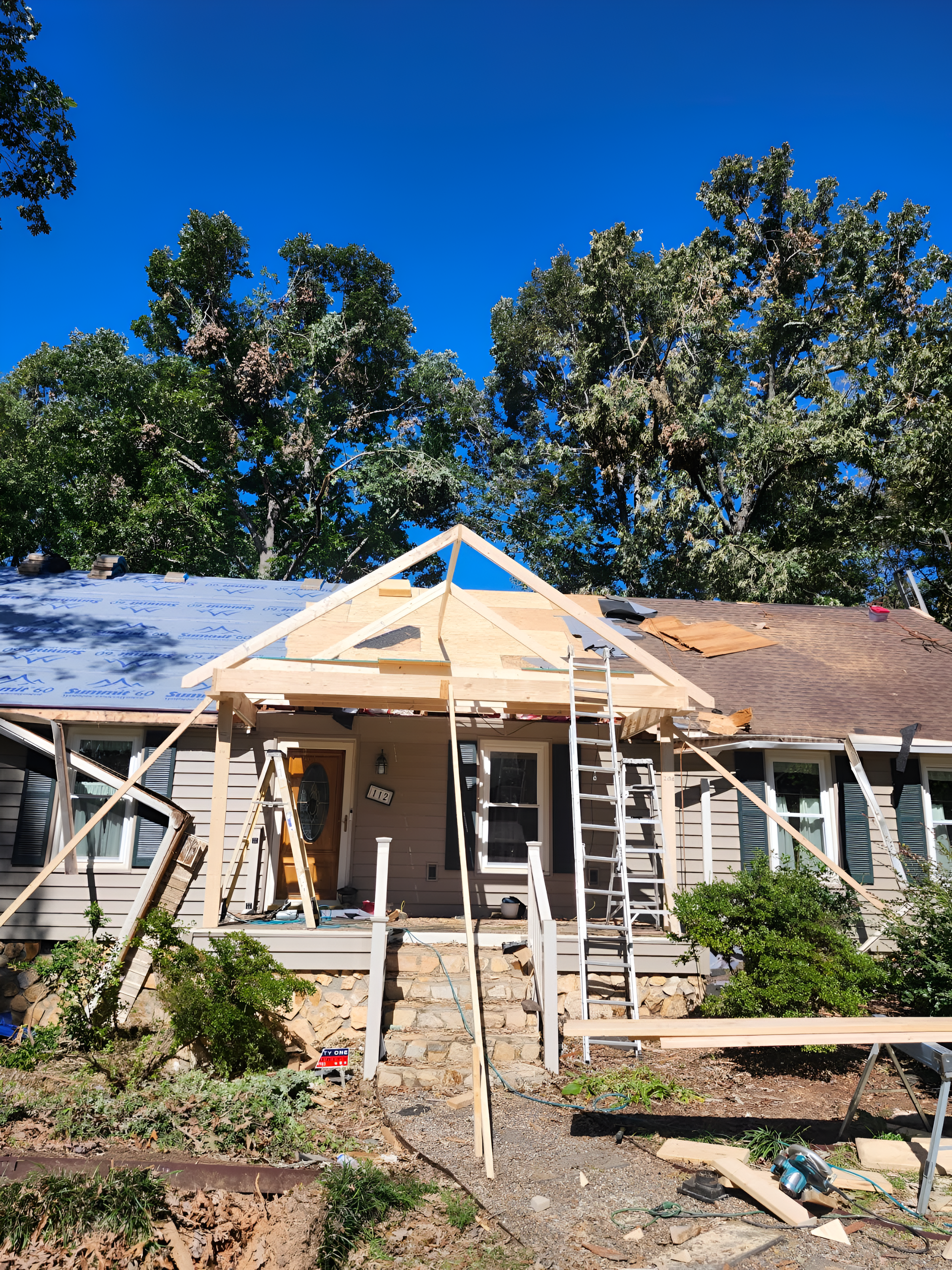 House under construction with a new wooden porch roof. Ladder and scaffolding in use; clear blue sky.