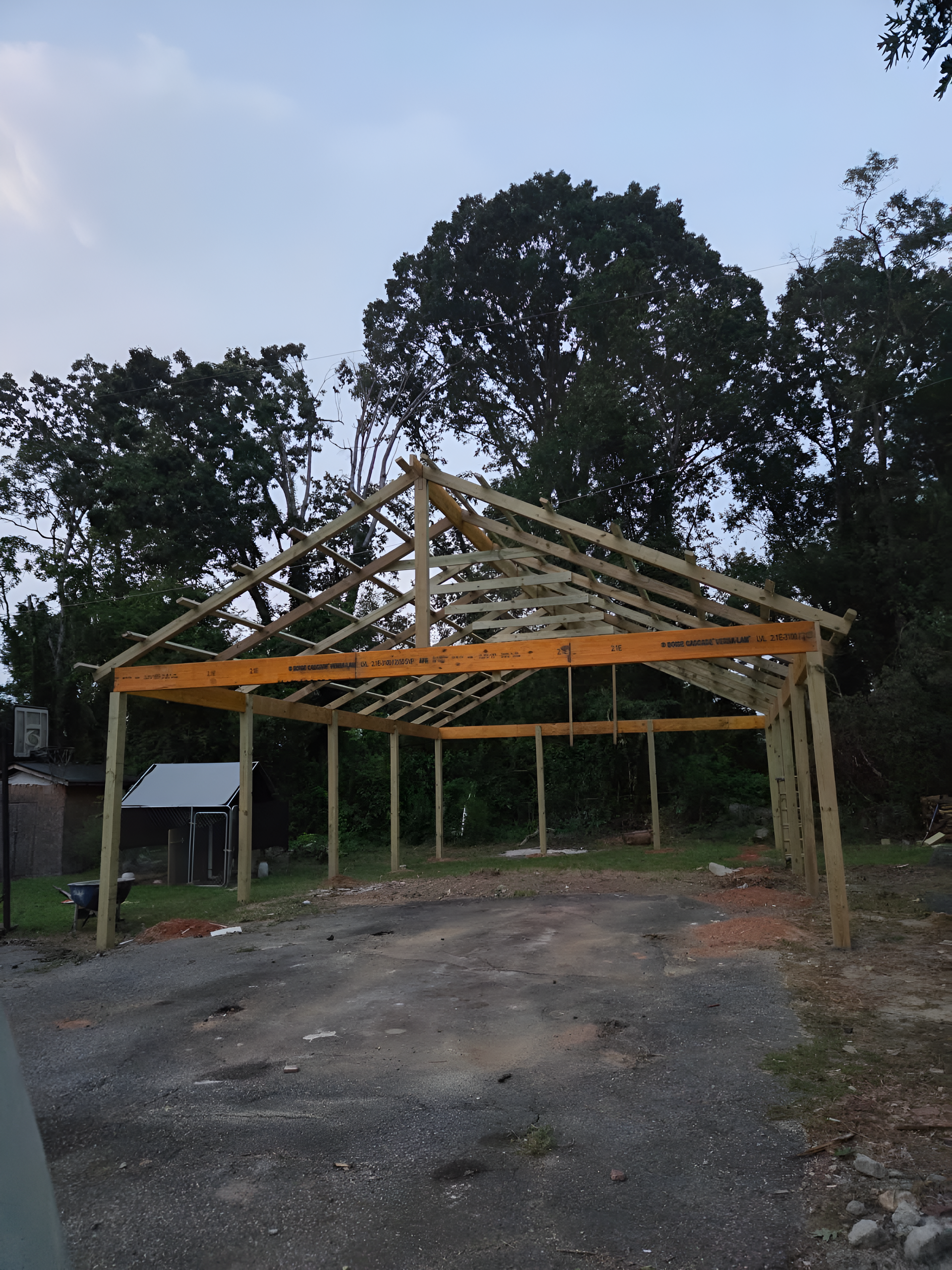 Wooden carport under construction in a gravel driveway, with trees in background.