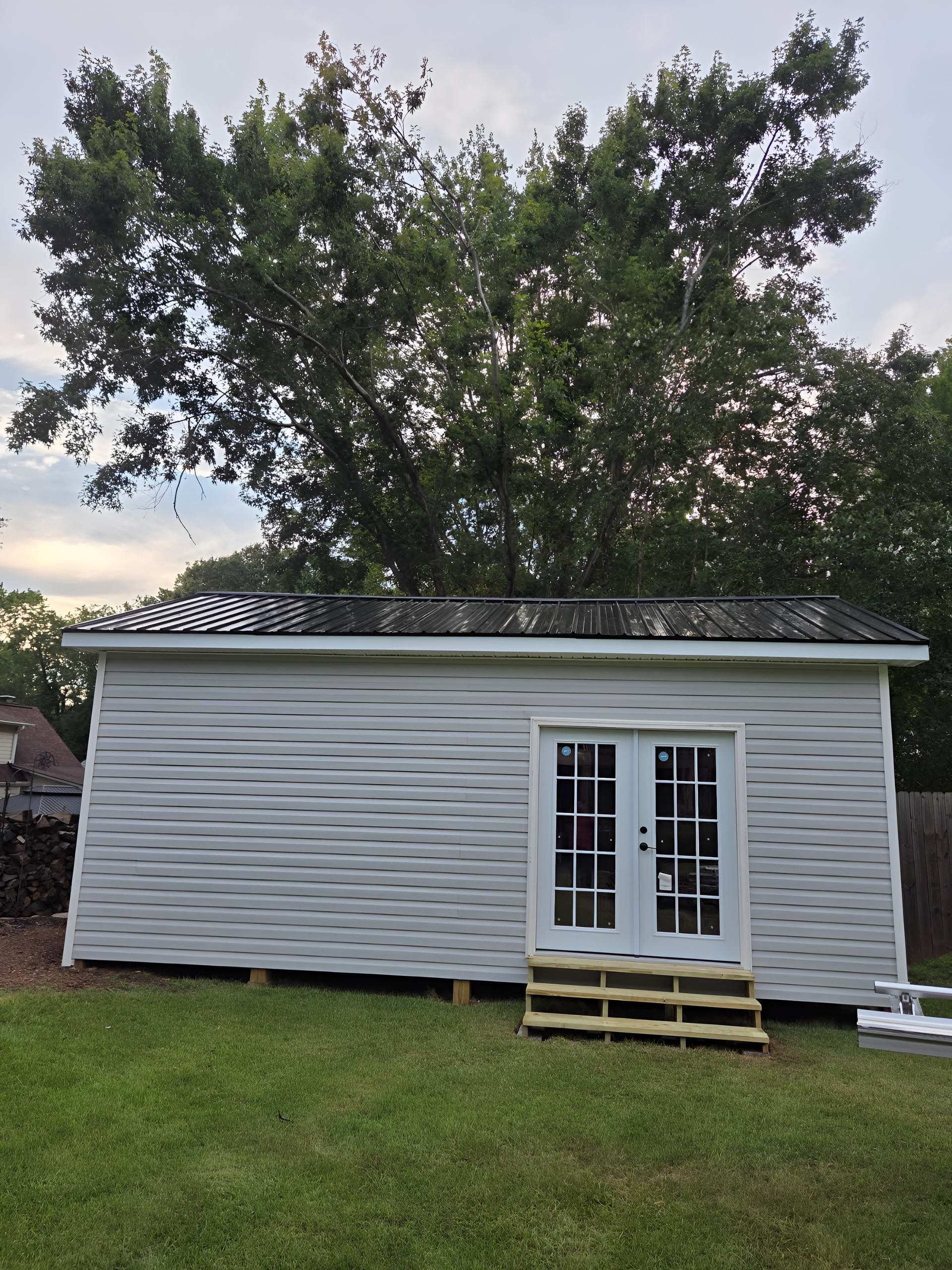White shed with French doors and wood steps in a grassy backyard. Tree in the background.