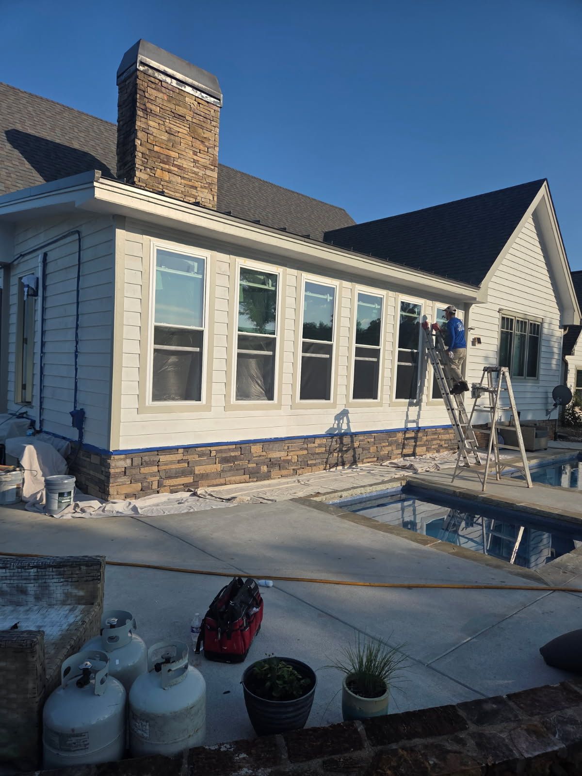 Exterior of a house being painted near a pool. A worker on a ladder. White siding with stone accents.