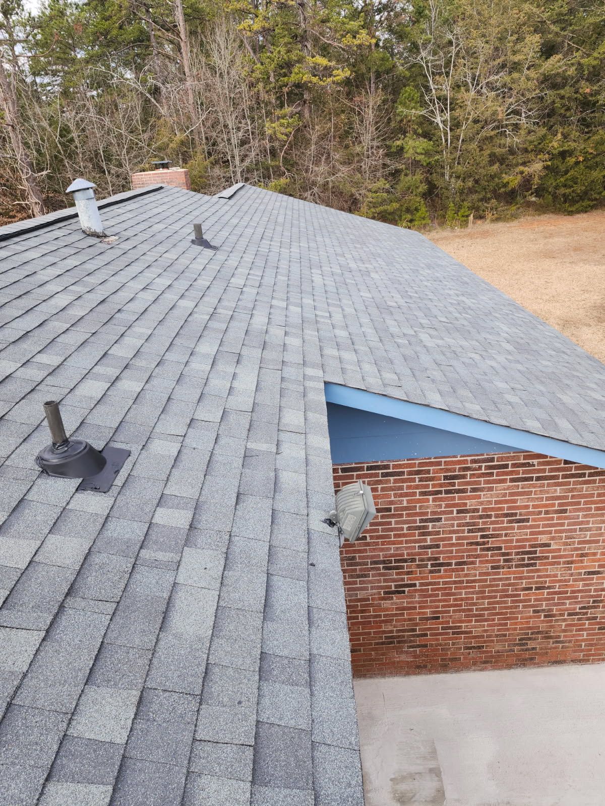 Gray shingle roof of a house with vents, near a red brick wall and trees.