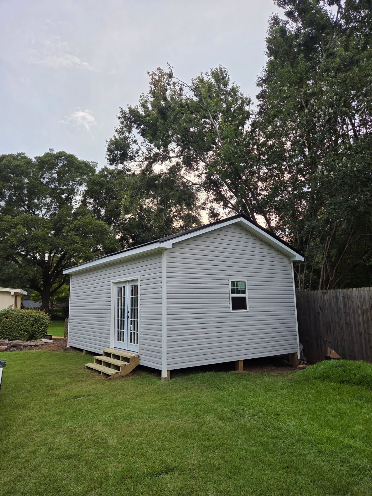 A small, light gray shed with a black roof and white double doors, set on a green lawn.