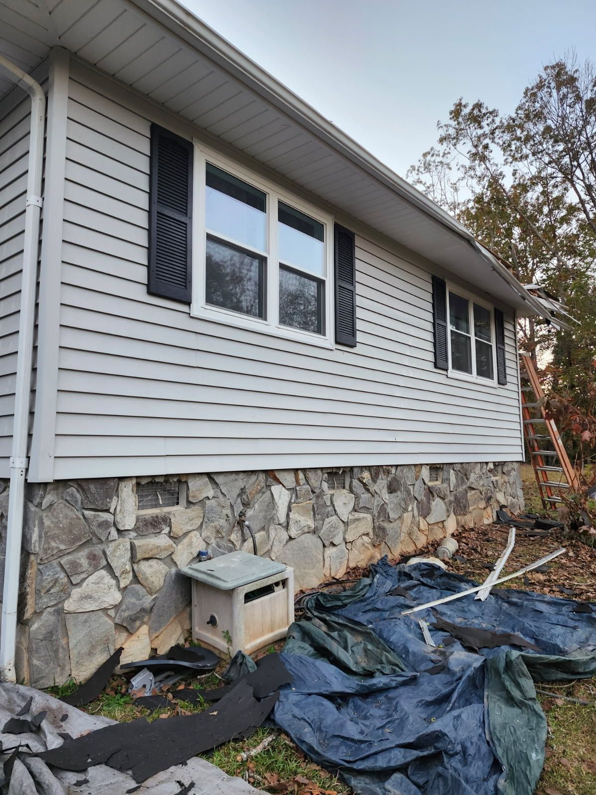 A house with light siding, dark shutters, and a stone foundation; a ladder is to the right.
