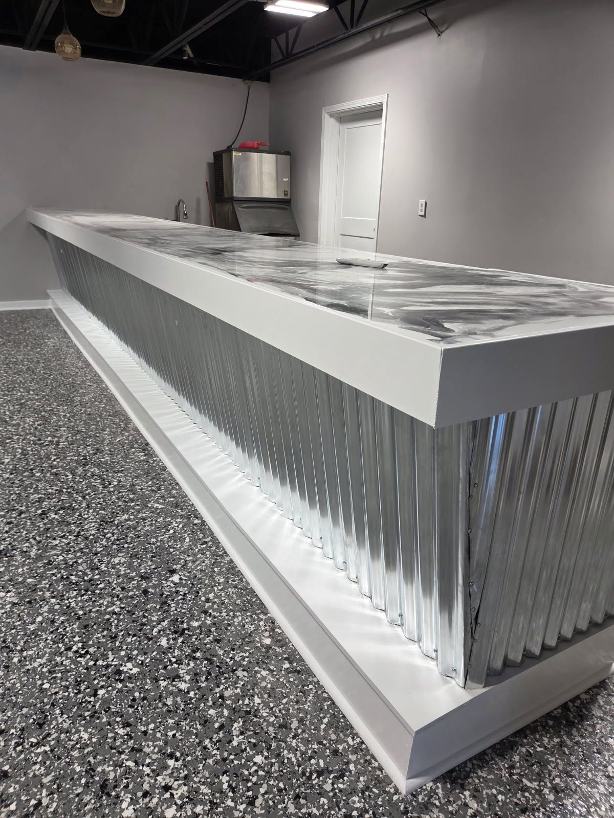 Modern bar with corrugated metal sides, white trim, and a marbled countertop, in a room with a speckled floor.