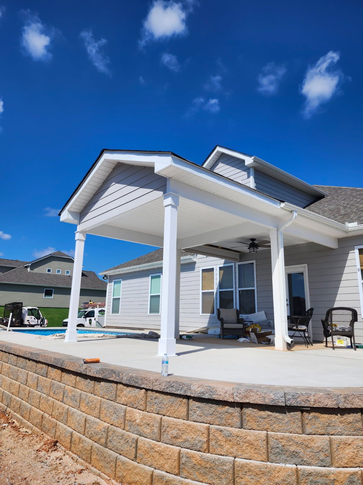 Covered patio attached to a house with white pillars, grey siding, and a blue sky.
