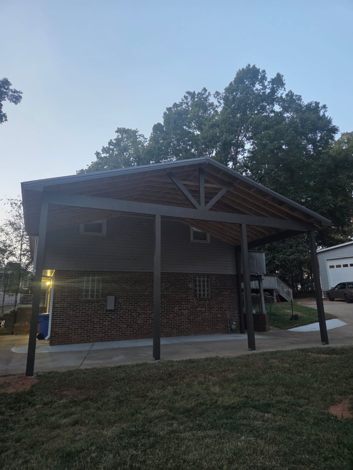 Two-story structure with a carport. Dark wooden frame, brick facade. Trees and car in background.