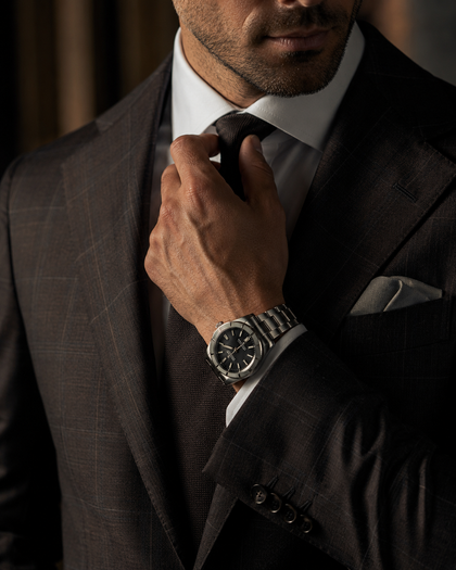 Un uomo in abito scuro si sistema la cravatta, indossa un orologio d'argento e un fazzoletto da taschino.