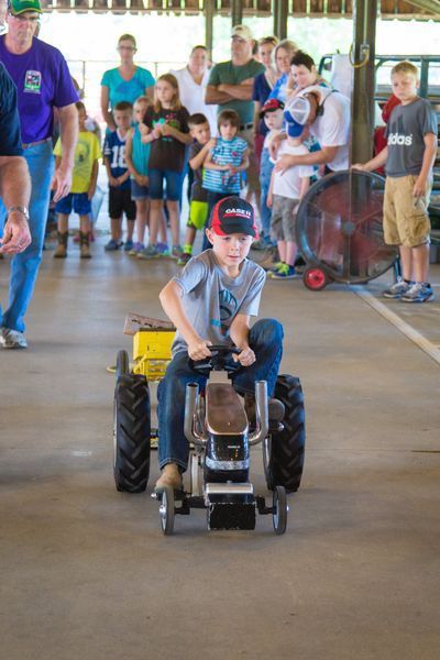 Child driving a small tractor indoors while people watch in the background