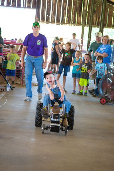 Child driving toy car in a barn while a group of people watch nearby