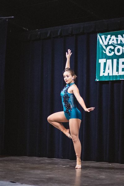 Young dancer in a blue costume striking a pose onstage with one arm raised.