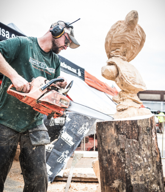Chainsaw carver shaping a wooden owl sculpture on a stump outdoors.