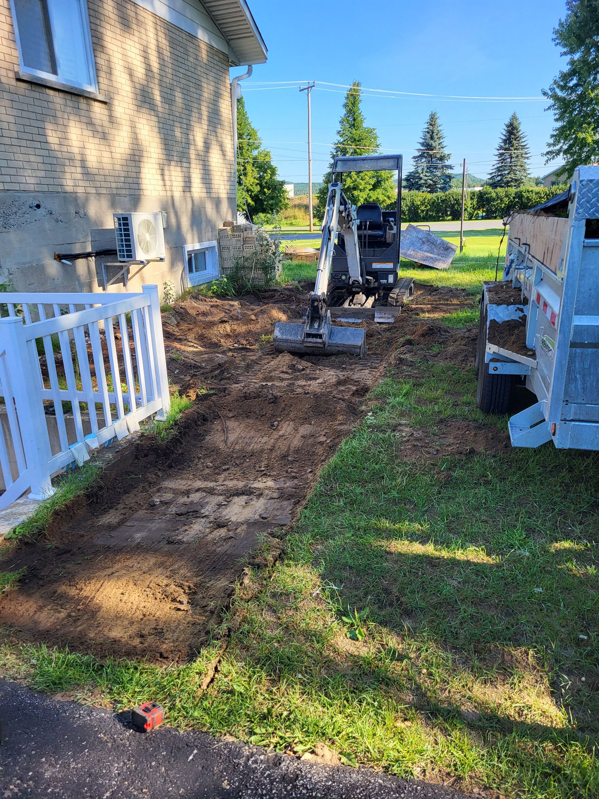 Une petite excavatrice creuse un chemin devant une maison.