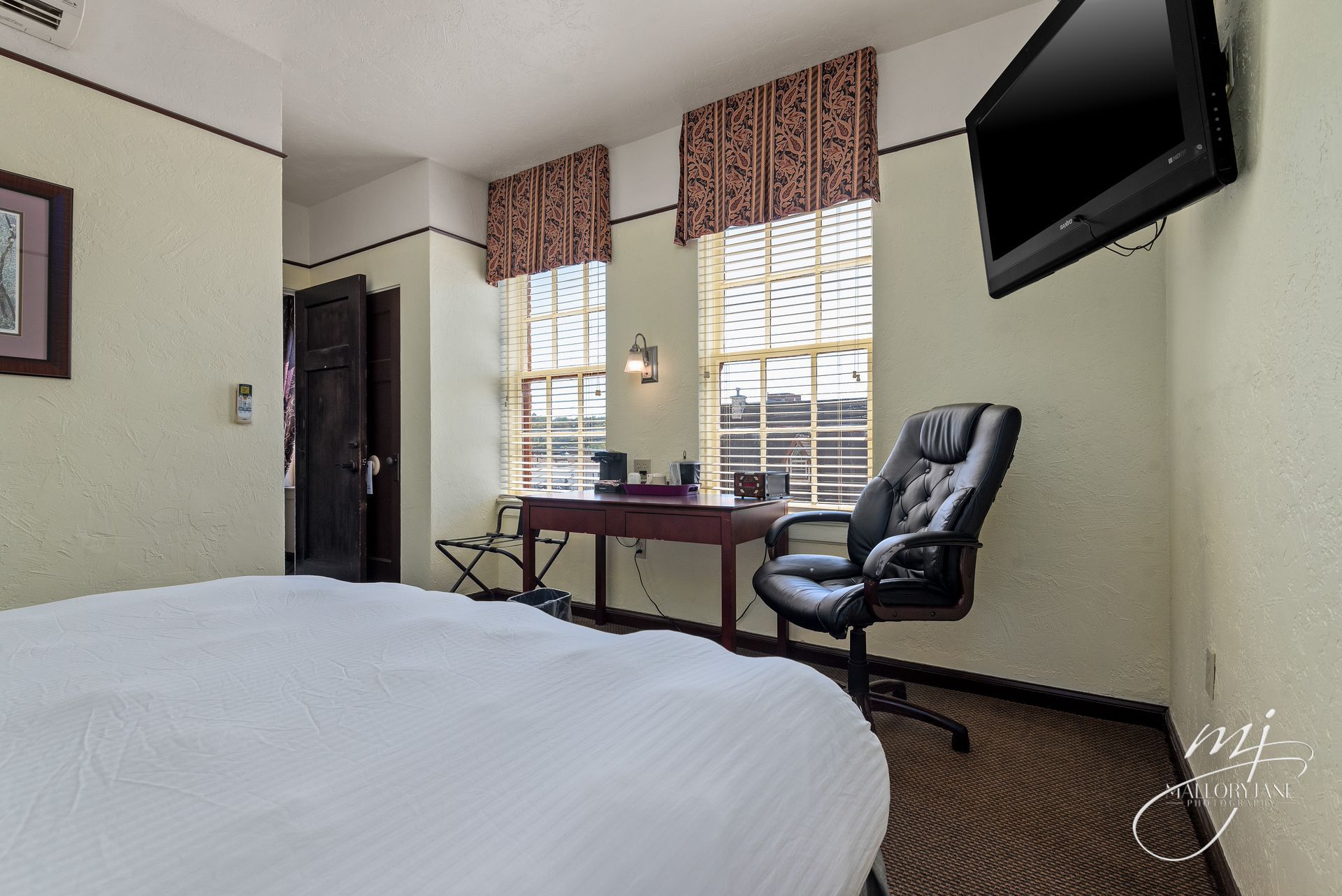 Hotel room with desk, TV, bed, two windows with blinds, and black leather office chair.