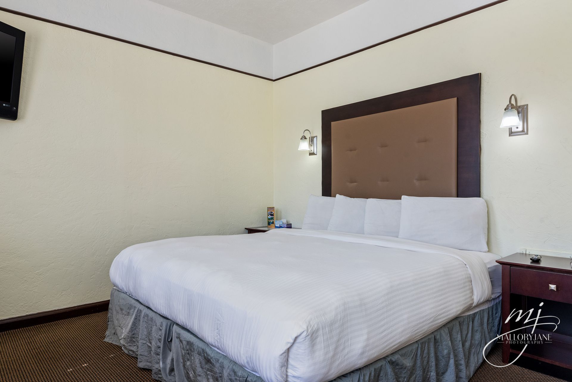 Hotel room with a bed, headboard, lamps, and a TV on the wall. Beige walls and brown furniture.