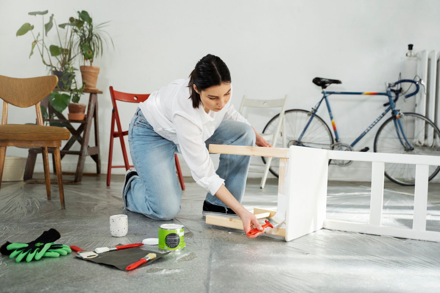 A woman is kneeling on the floor painting a bench in a living room.