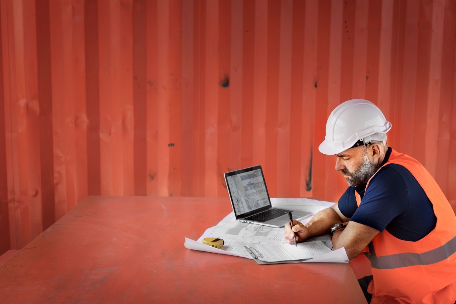 A construction worker is sitting at a table with a laptop and a blueprint.