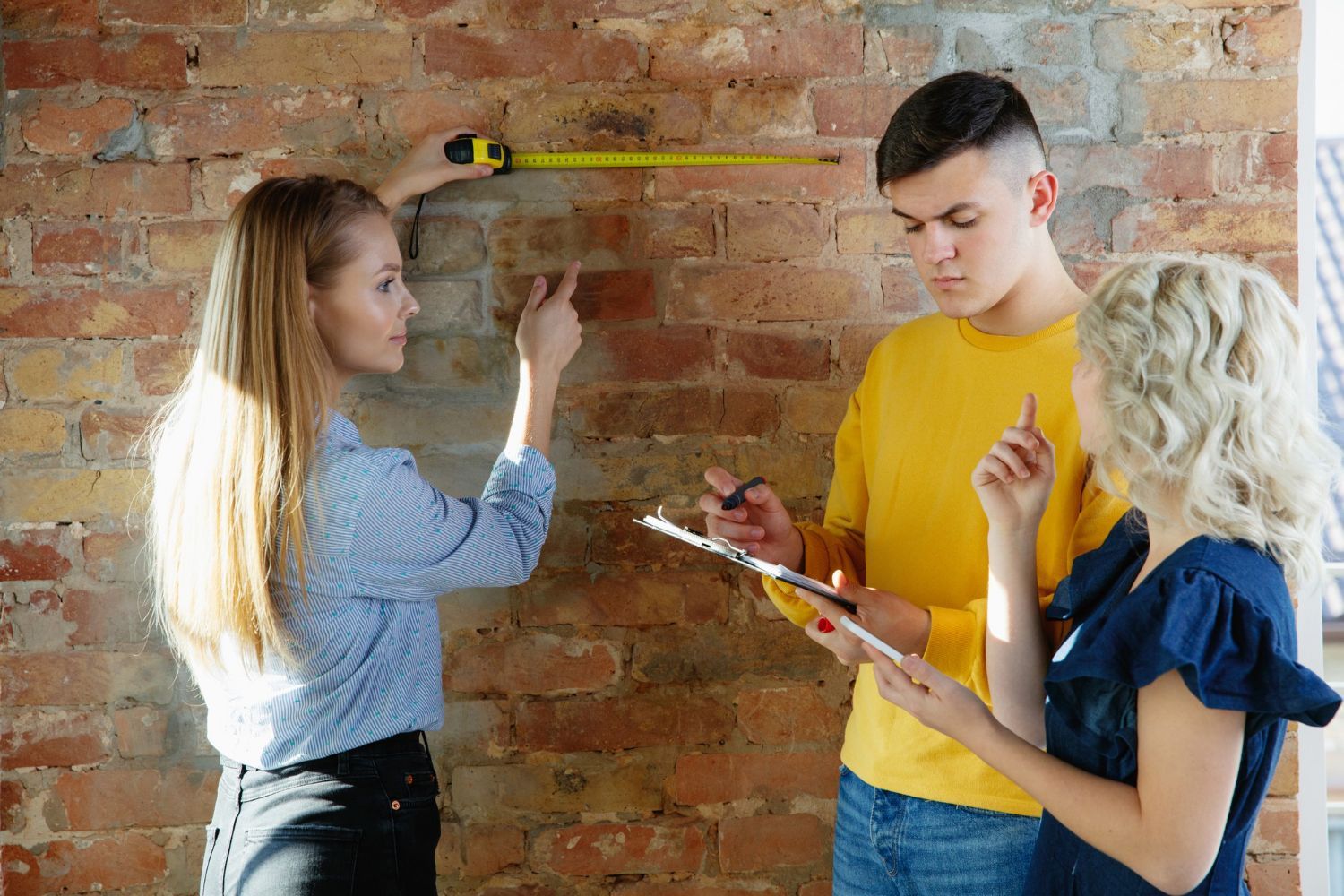 A man and two women are measuring a brick wall with a tape measure.