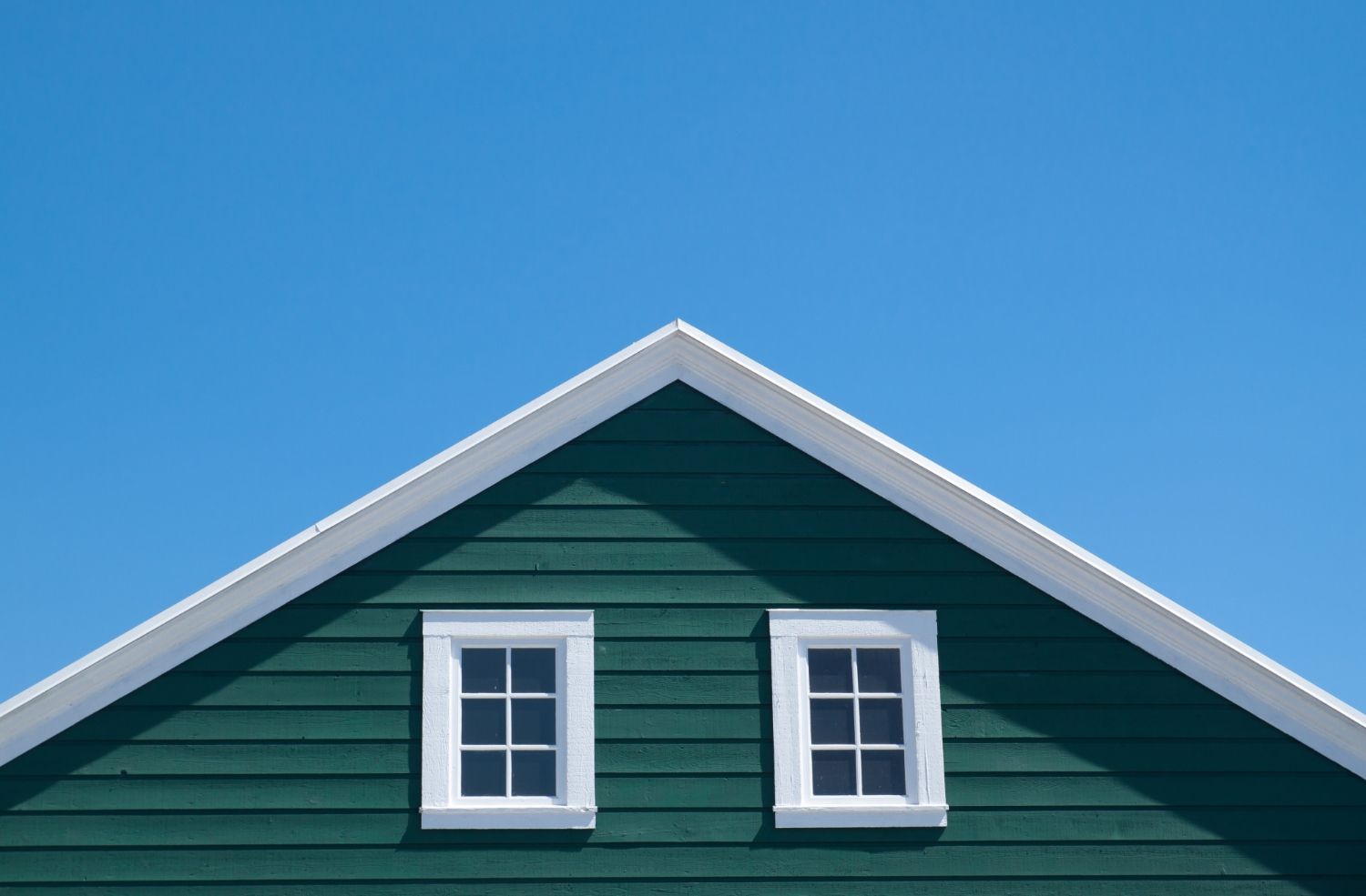 The roof of a green house with two windows against a blue sky.