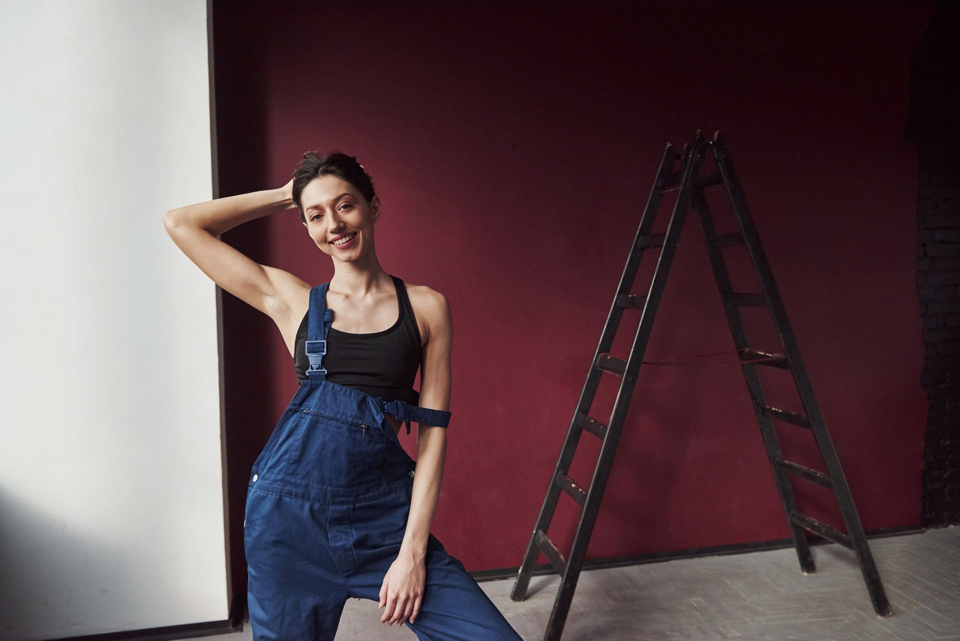 A woman in overalls is standing next to a ladder in a room.